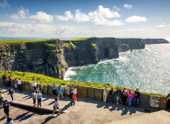 Image of the Cliffs of Moher on a bright day with a turquoise blue sea and bright green grass. People in foreground standing on path